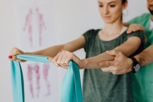Crop anonymous Physiotherapist in wristwatch and uniform helping young woman in casual wear reaching arms with elastic tape in doctor office