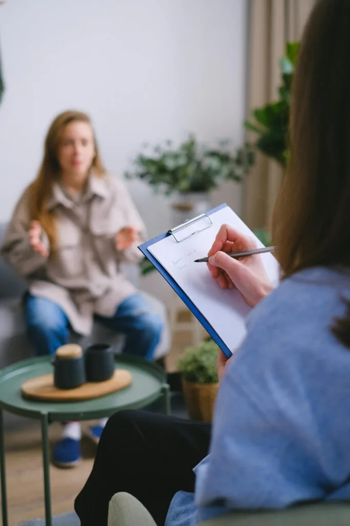 Female psychologist taking notes on clipboard while listening to patient sitting on blurred background during psychotherapy consultation in office