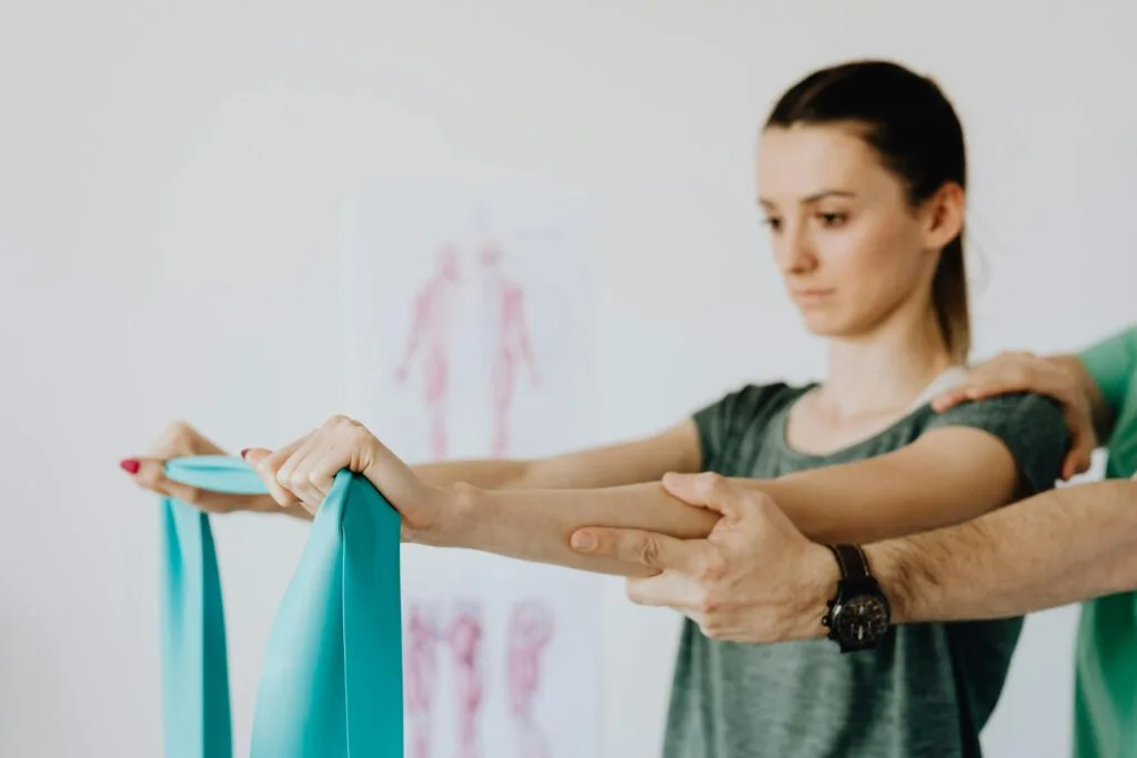 Slim young female in casual wear standing with reached arms and elastic tape during checkup by a StretchMasters® therapist in wristwatch