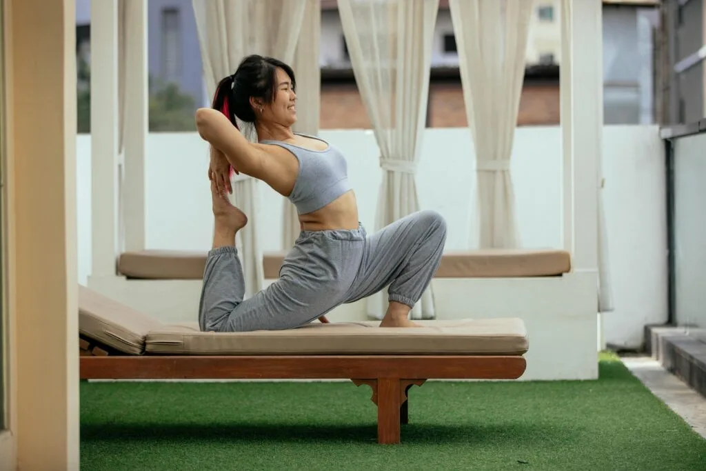 Side view of cheerful ethnic female stretching legs and practicing yoga in Crescent Lunge on the Knee position on deckchair on terrace