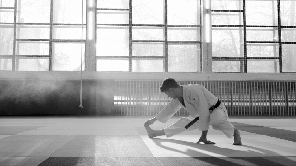 A martial artist in uniform stretches in a sunlit dojo, focused and determined.
